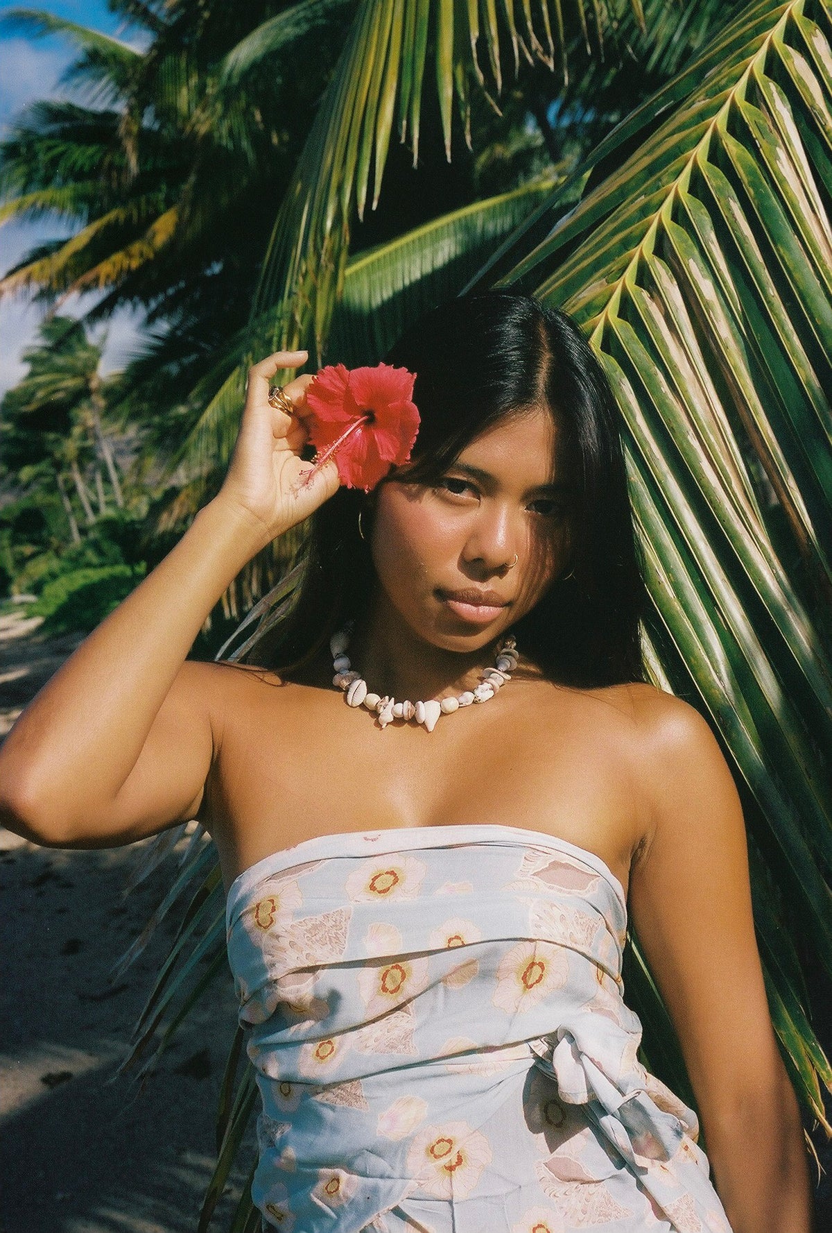 Woman in a strapless dress holding a red flower in front of her face, standing against a palm tree.