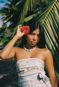 Woman in a strapless dress holding a red flower in front of her face, standing against a palm tree.