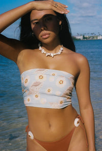 Woman in a floral bandeau top and brown bikini bottom standing by the water.