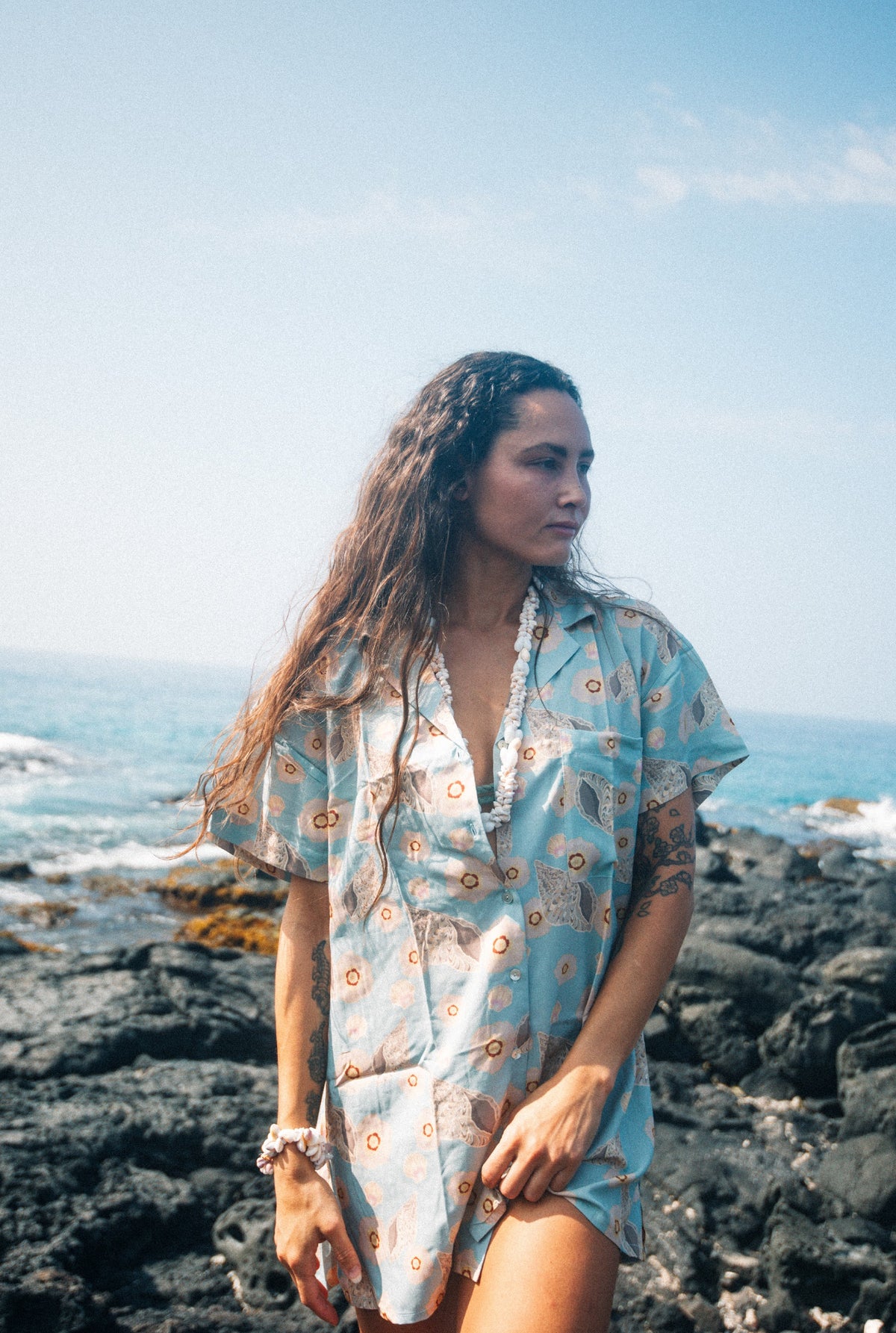 A person wearing a short-sleeved shirt dress with a pattern that includes images of shells and flowers on a baby blue background, standing on a rocky coast with the ocean and sky in the backdrop.