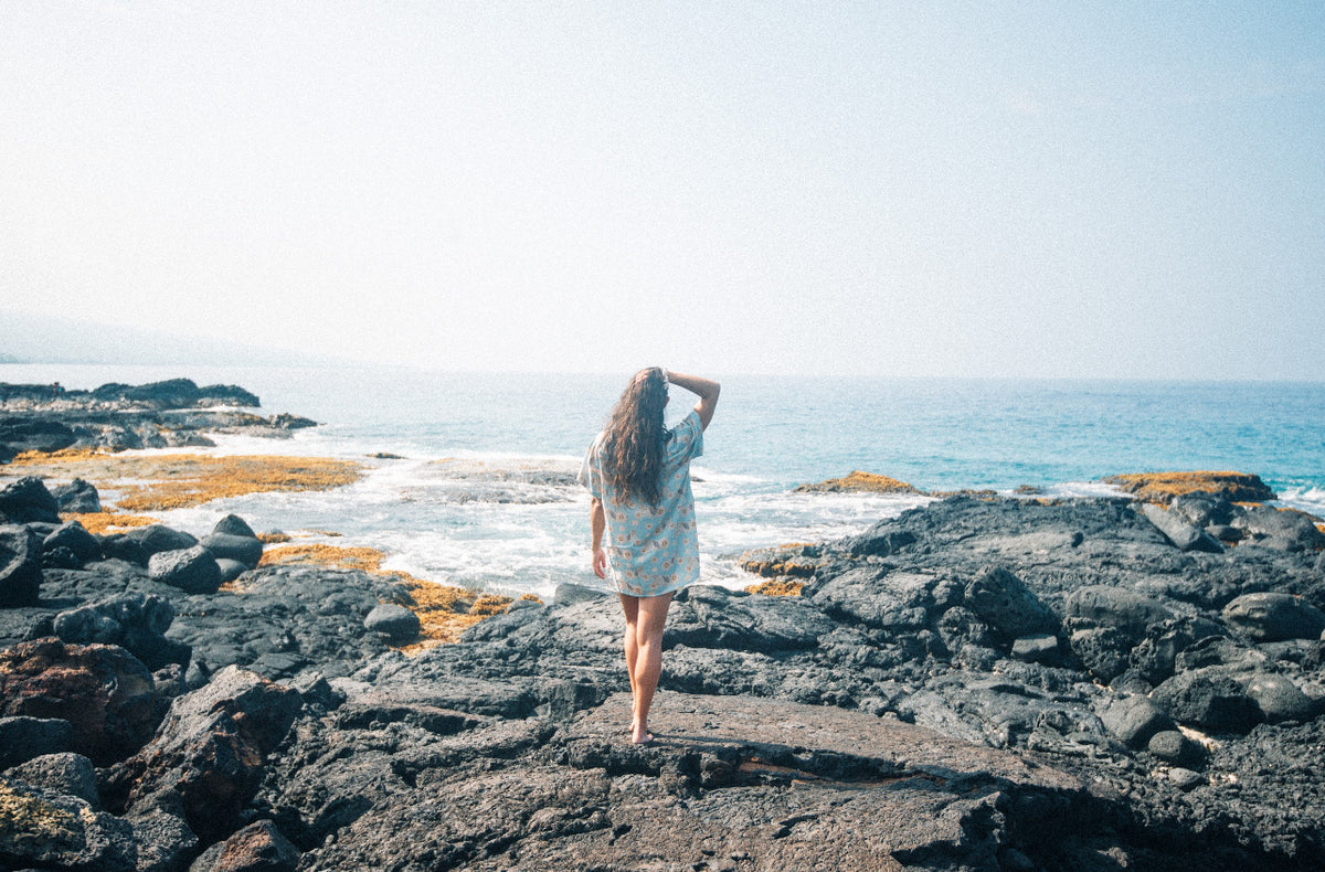 A person standing on a beach wearing a short-sleeved shirt dress with a floral pattern on a baby blue background.