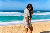 A person standing on a beach wearing a short-sleeved shirt dress with a floral pattern on a baby blue background.