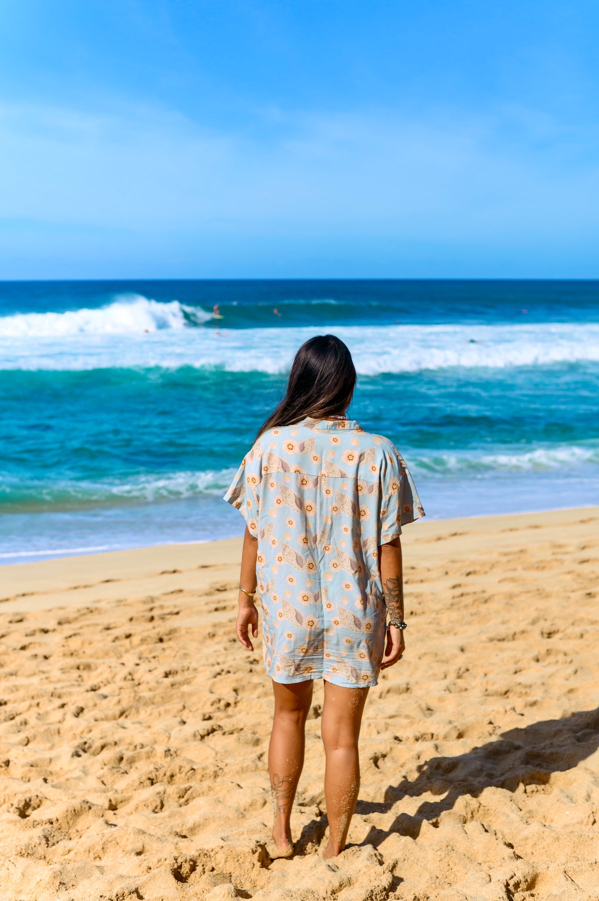 A person standing on a beach wearing a short-sleeved shirt dress with a floral pattern on a baby blue background.