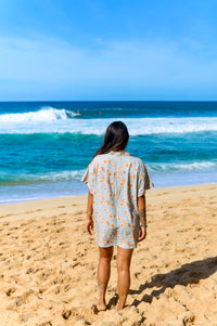 A person standing on a beach wearing a short-sleeved shirt dress with a floral pattern on a baby blue background.