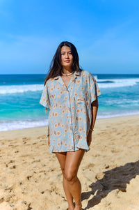 A person standing on a beach wearing a short-sleeved shirt dress with a floral pattern on a baby blue background.