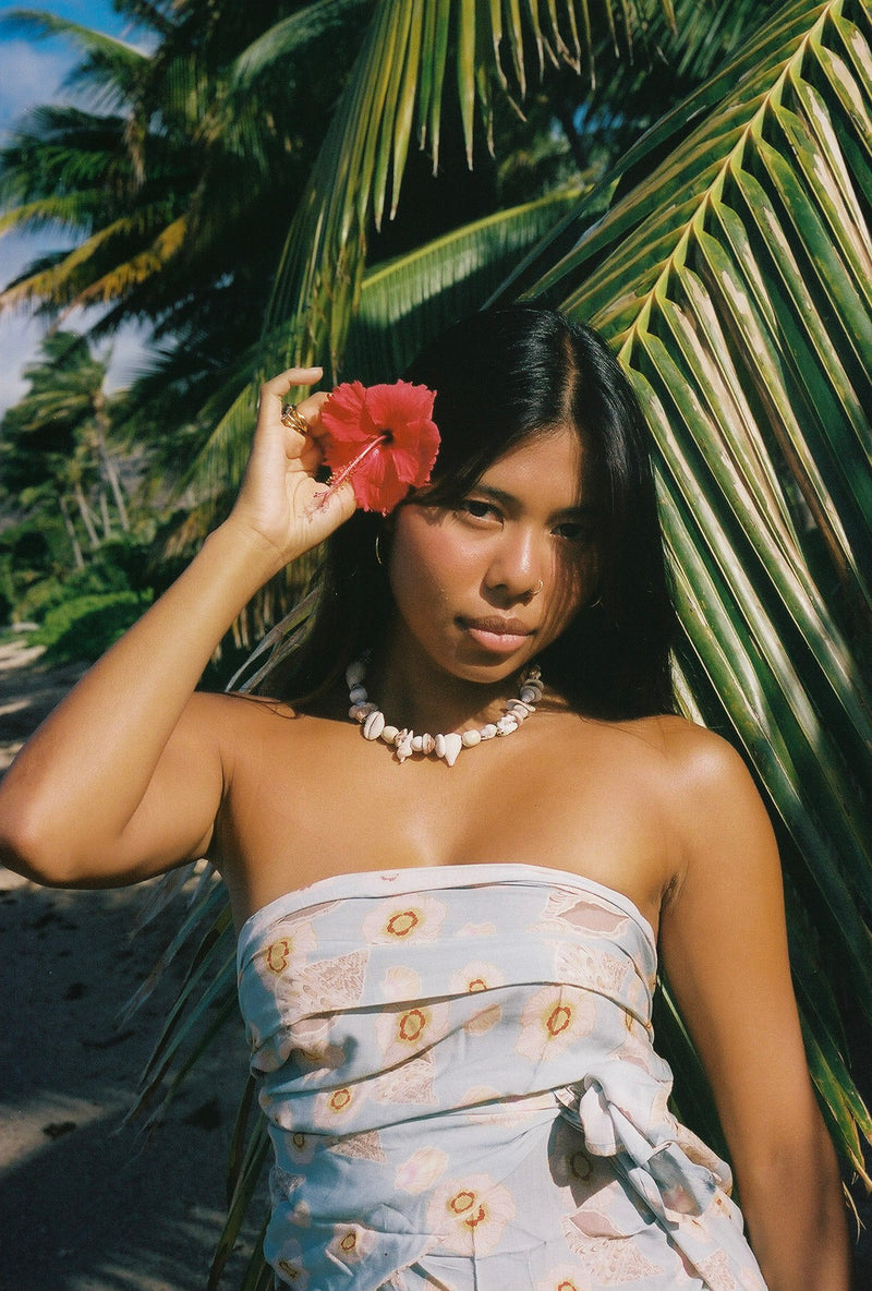 Woman in a strapless dress holding a red flower in front of her face, standing against a palm tree.