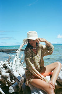 Woman sitting on driftwood by the ocean wearing a hat and light dress.