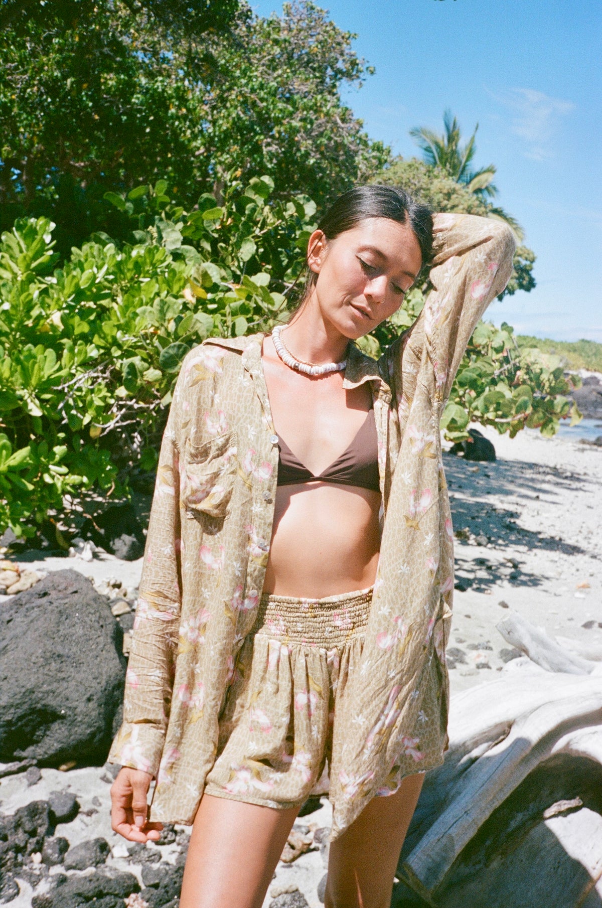 Woman in a bikini and cover-up standing on a rocky beach with greenery in the background
