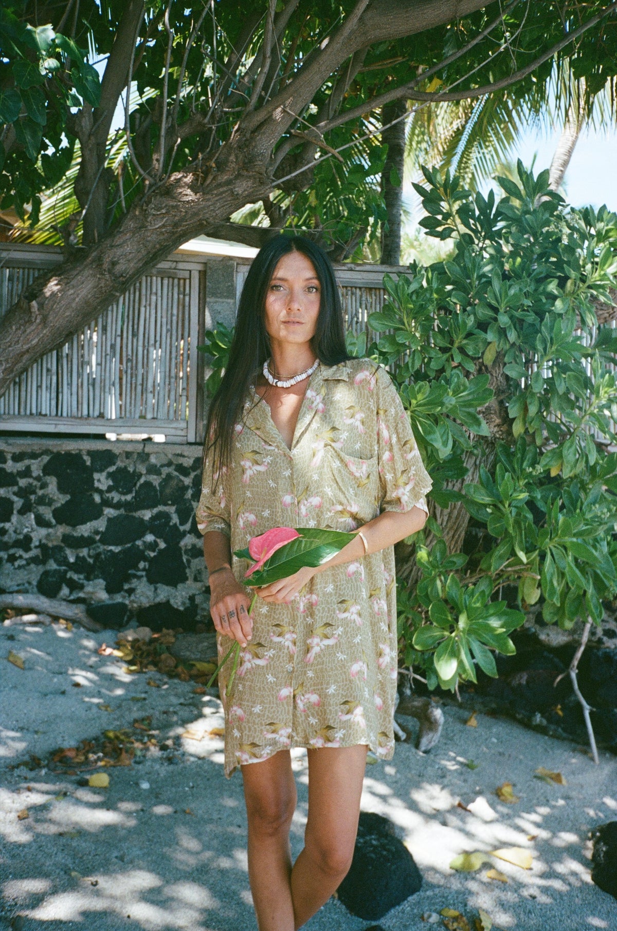 Woman in a floral dress standing outdoors with greenery and a building in the background