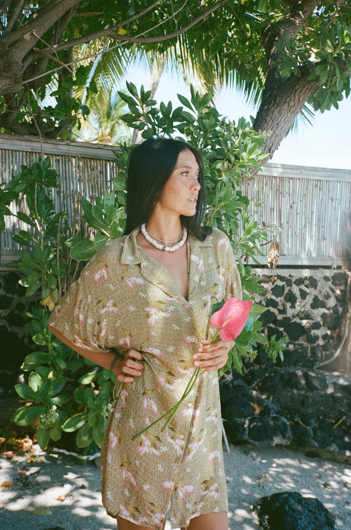 Woman in a patterned dress holding a pink flower in a tropical setting