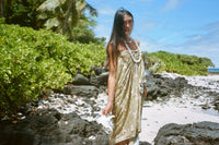 Woman in a dress standing on a rocky beach with greenery and blue sky.
