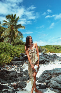 Woman in a dress standing on a rocky beach with palm trees and blue sky.