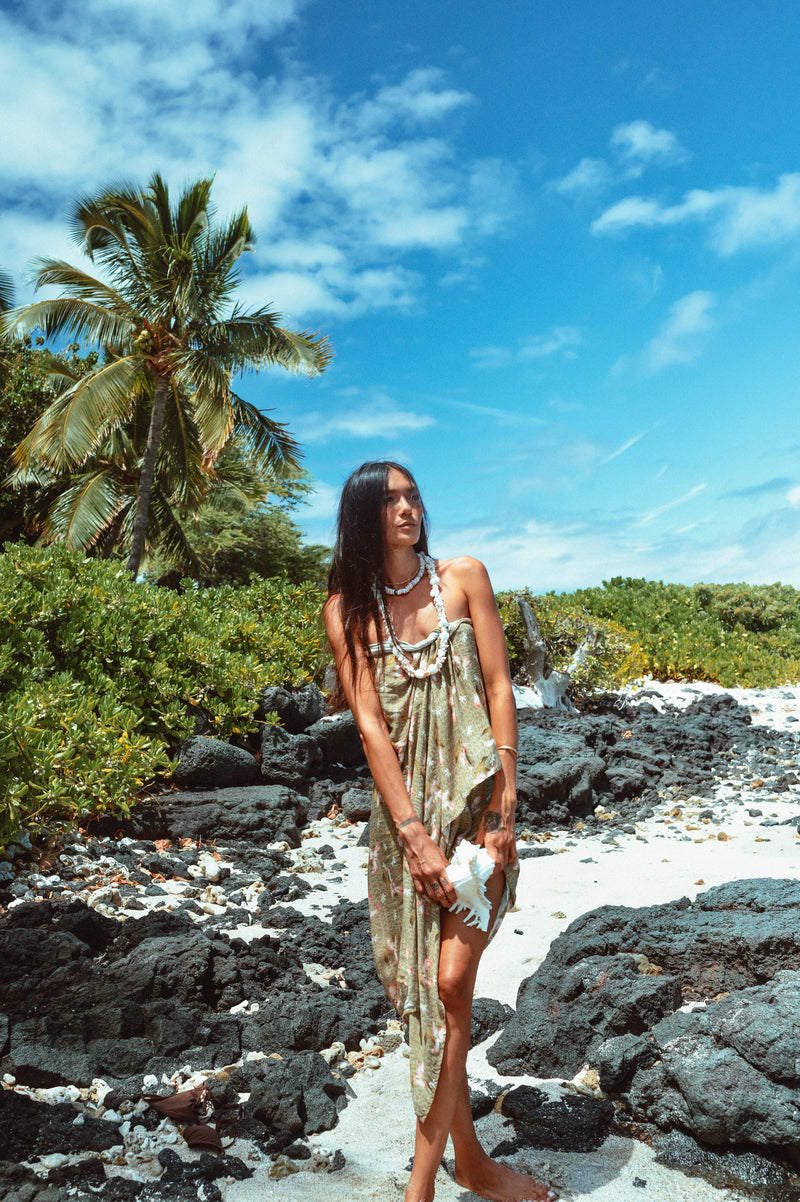 Woman in a dress standing on a rocky beach with palm trees and blue sky.