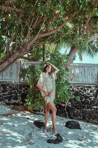 Woman in a white dress standing under a tree on a sandy path with a stone wall and greenery in the background.