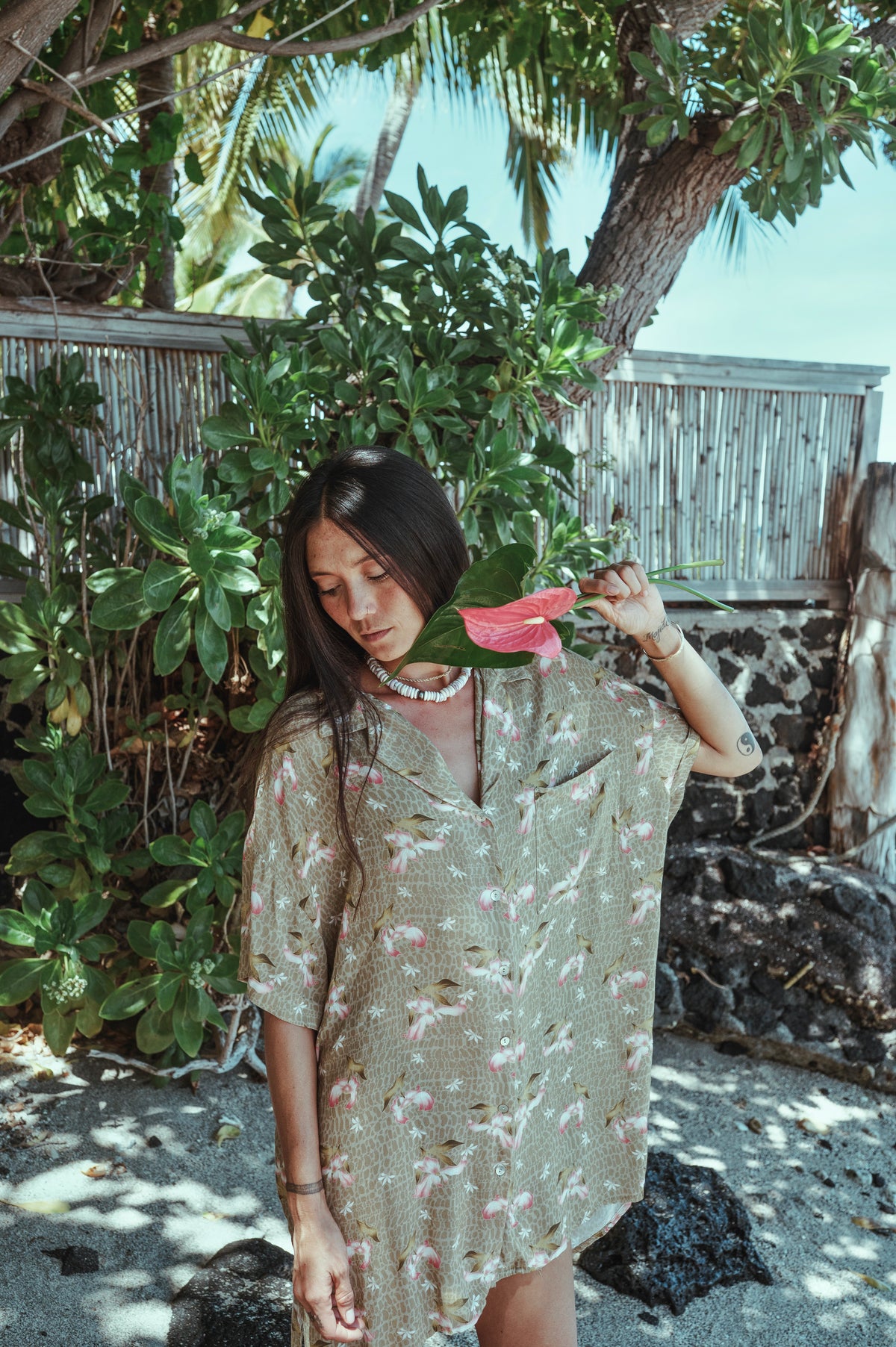 Woman holding a pink flower in a natural setting with greenery and wooden structures.