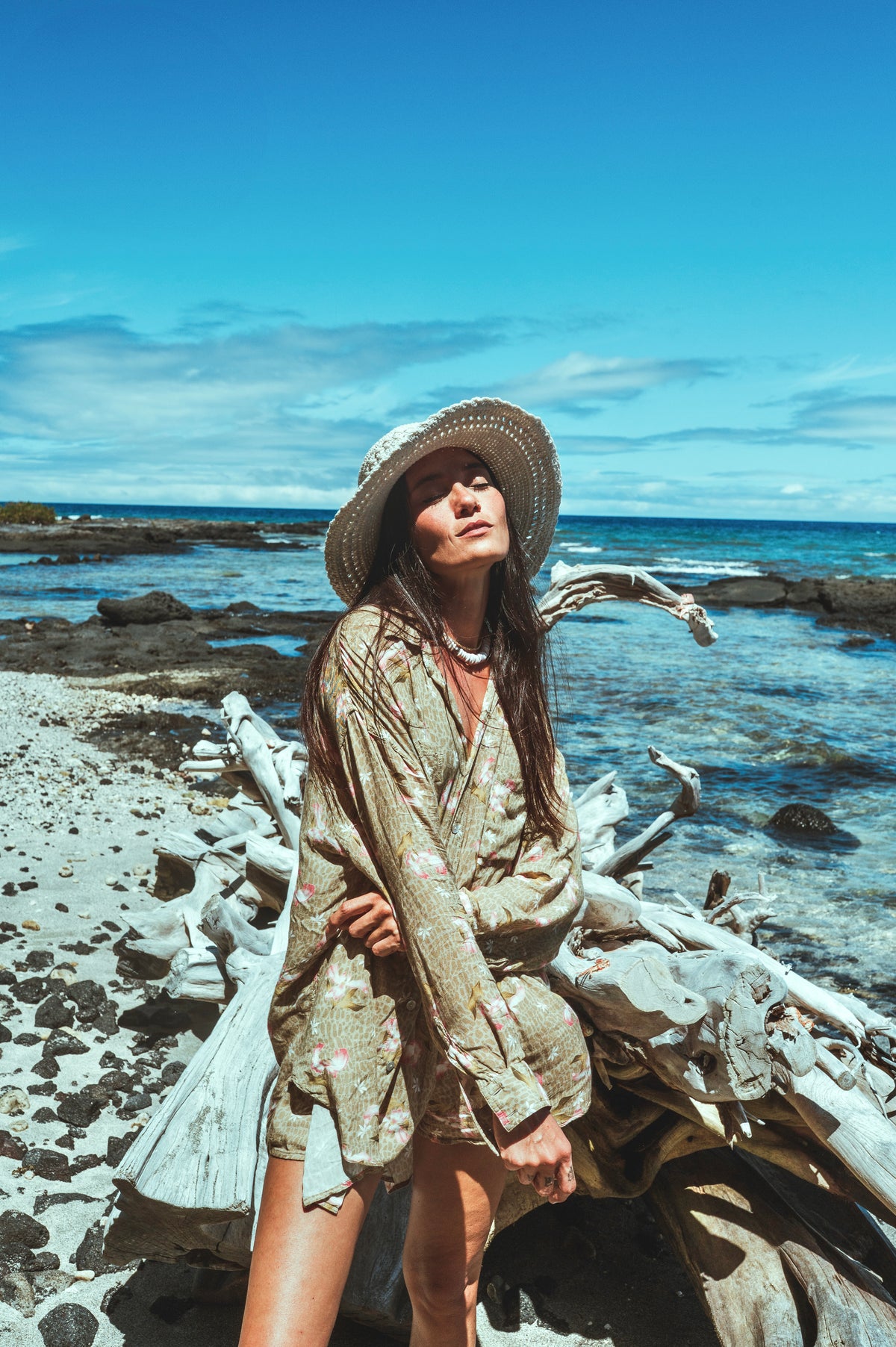 Woman in a straw hat and light outfit standing on a rocky beach with ocean and sky in the background