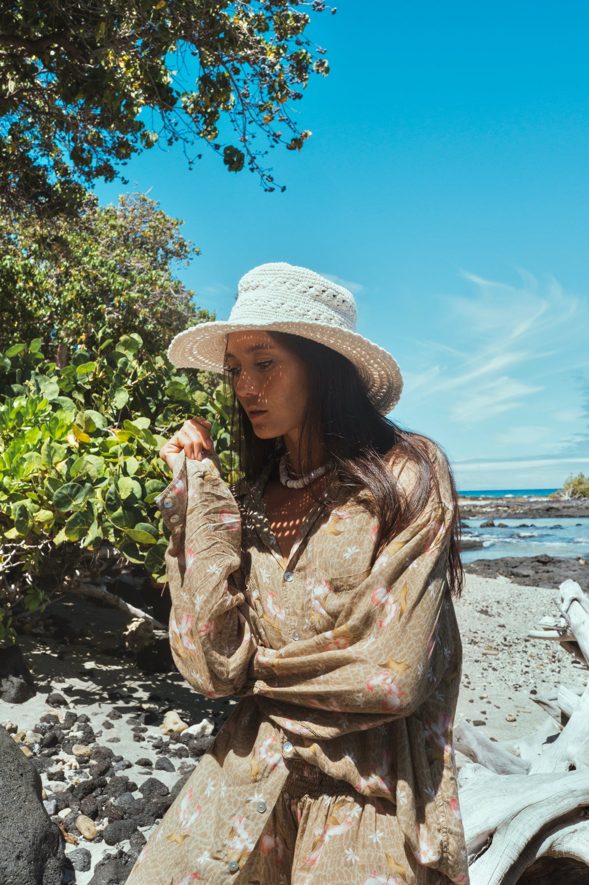 Woman in a straw hat and patterned dress standing on a rocky beach with greenery and blue sky.