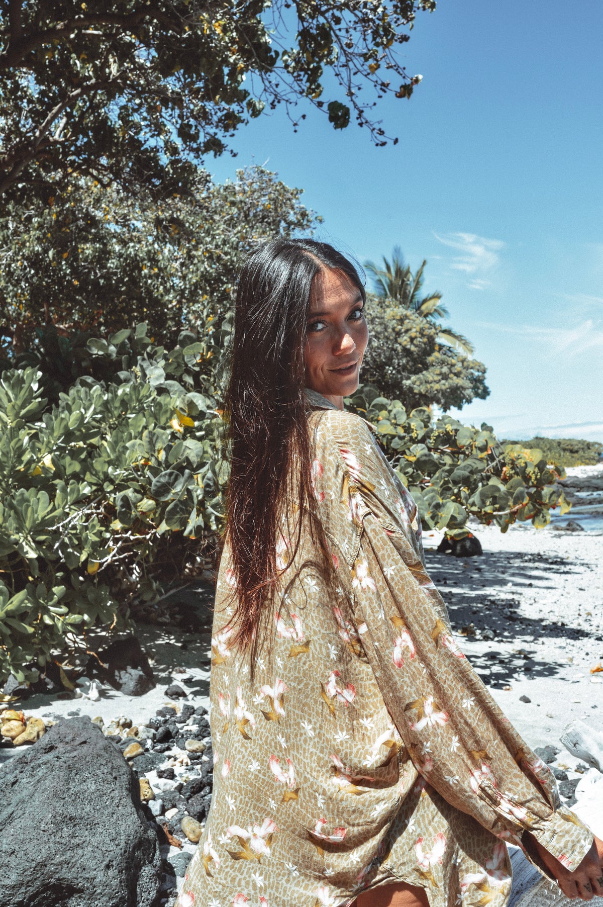 Woman in a floral dress standing on a beach with greenery and blue sky.