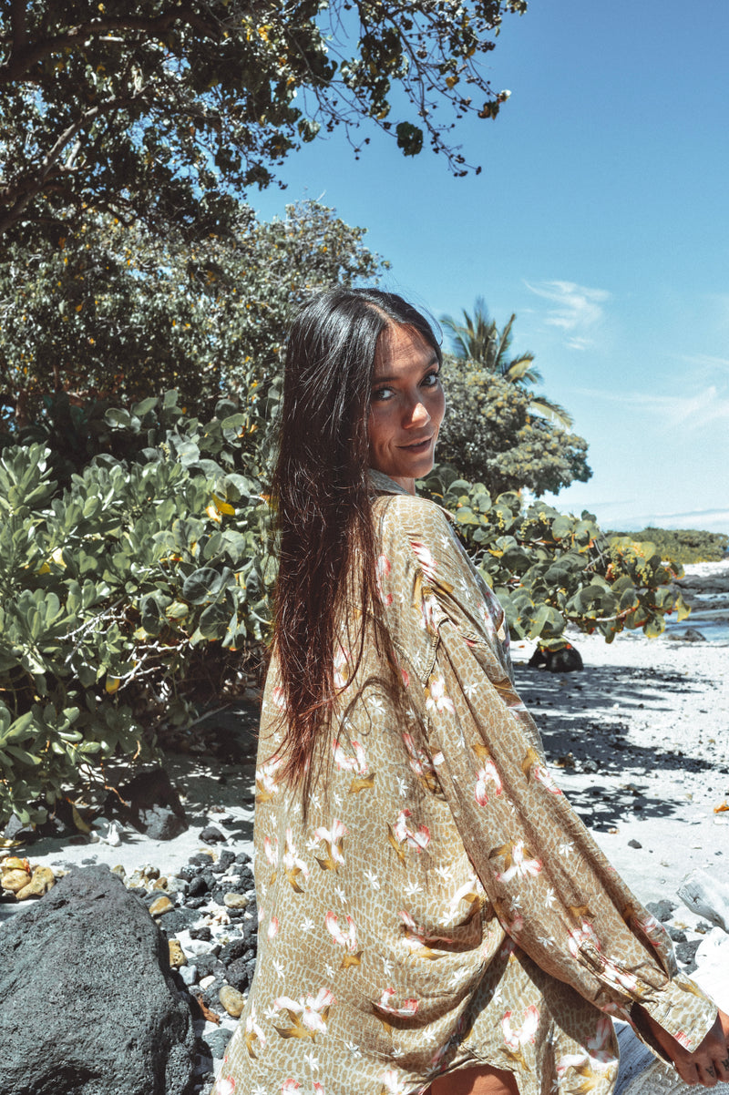 Woman in a floral dress standing on a beach with greenery and blue sky.