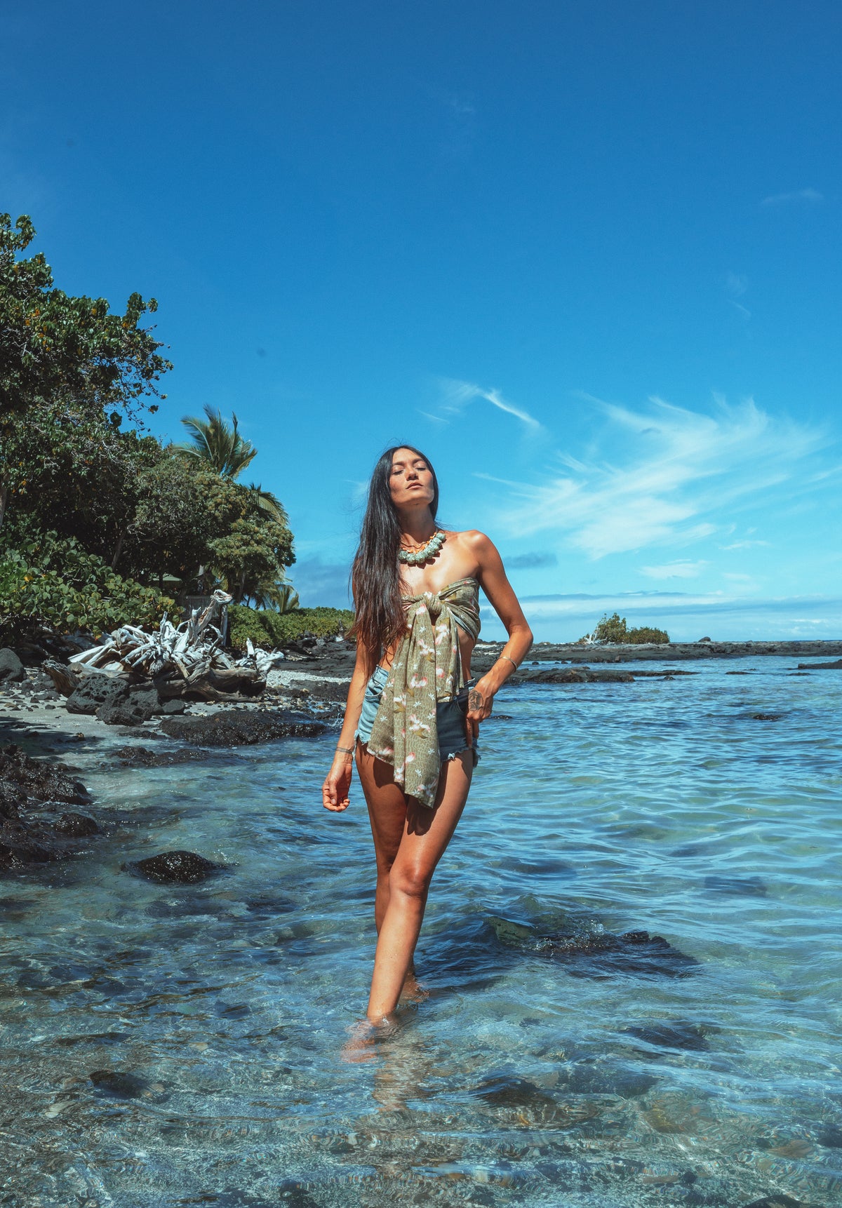 Woman walking in clear water near a tropical beach with blue sky and greenery.