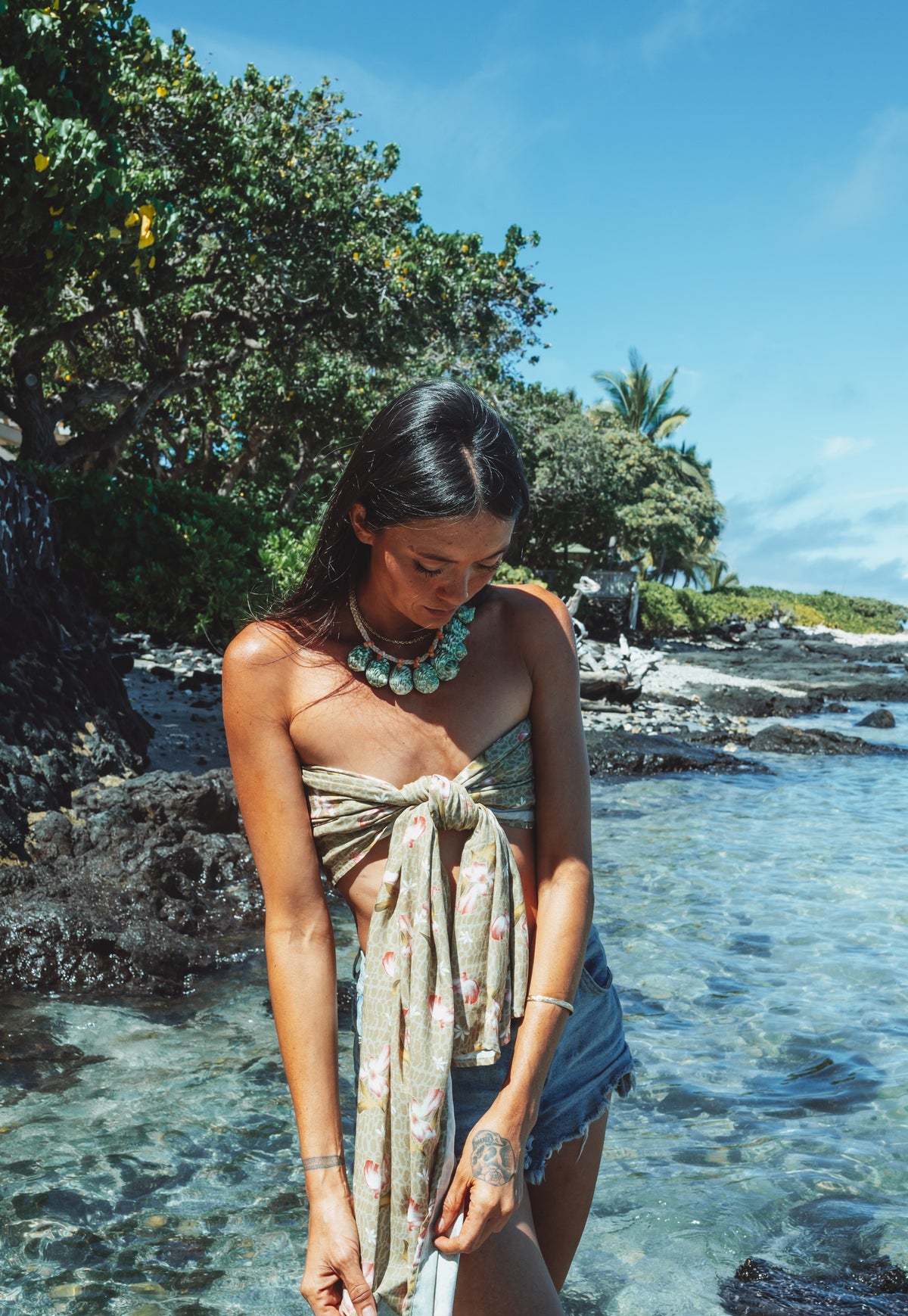 Woman standing by a rocky shoreline with clear blue water and greenery.